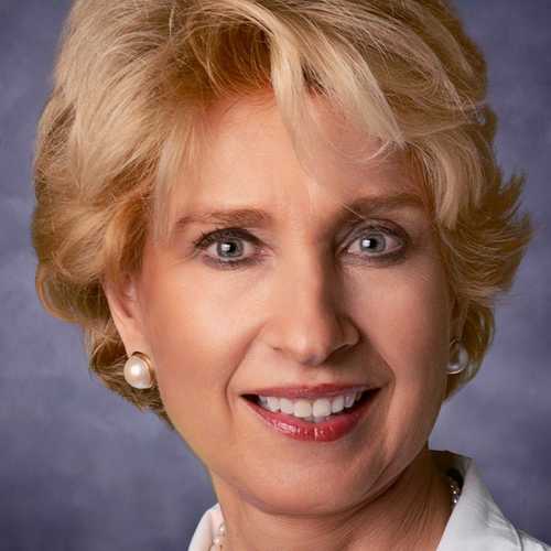 a blonde woman wearing a white lab coat smiles while looking into the camera