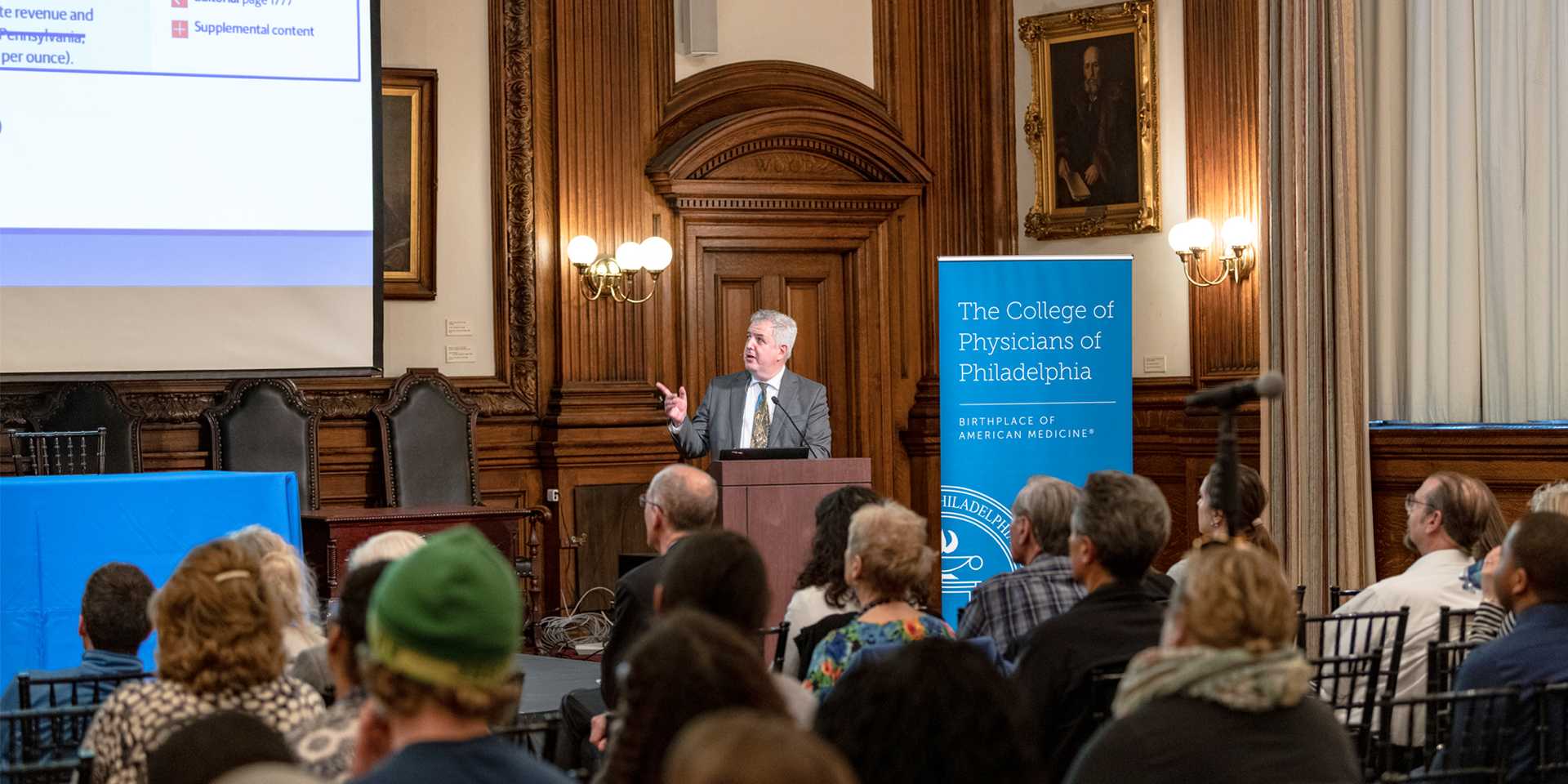 Speaker standing at a podium in front of a crowd and pointing towards a slideshow presentation