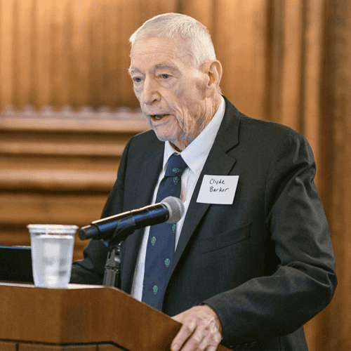Photo of Clyde Barker standing at a wooden podium in a dark suite with a blue tie